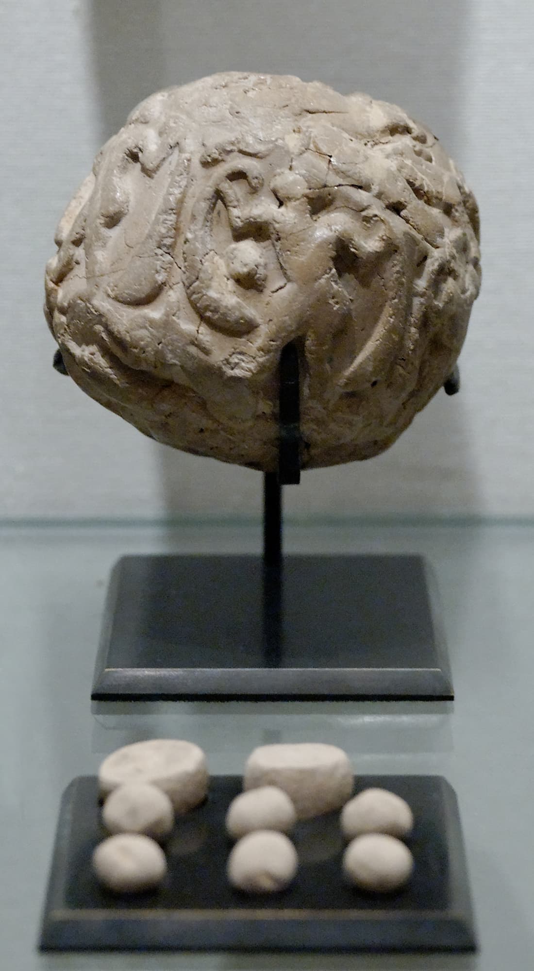 A round, rough clay ball on a stand, with eight small clay tokens of different shapes displayed below it.