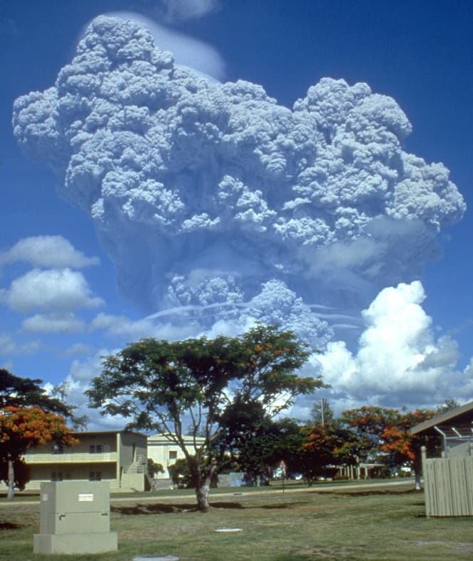 A huge grey eruption column rising above hills, seen from a distance under a blue sky.