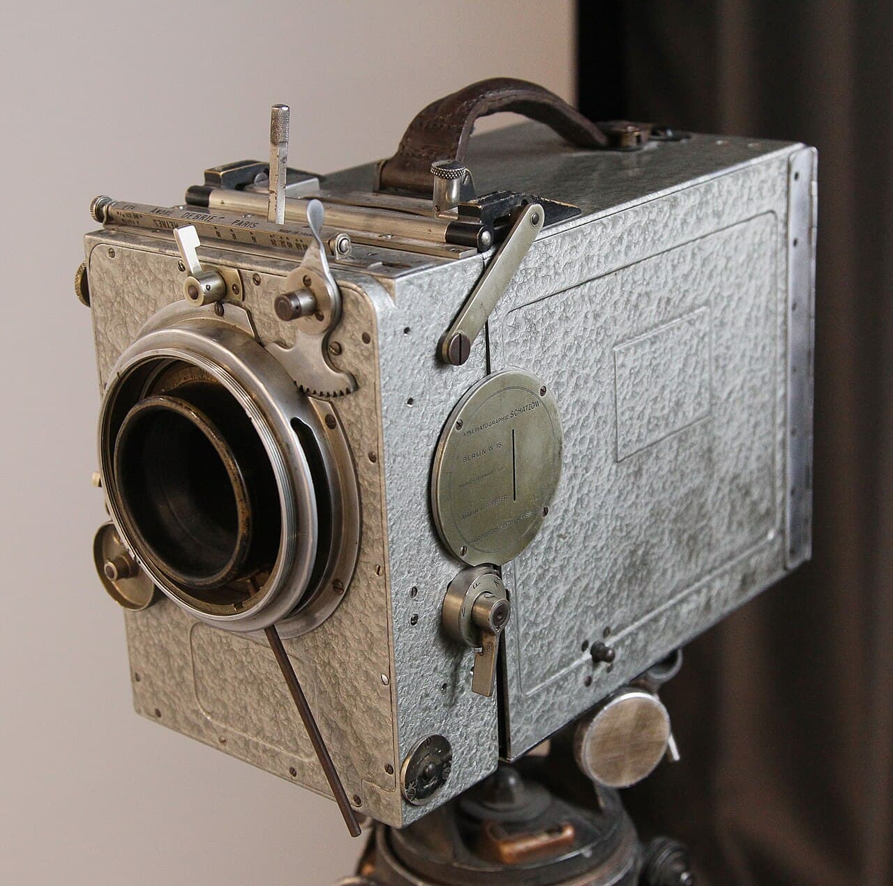 A wooden box-shaped Debrie Parvo movie camera with a lens on the front and a hand crank on its side, on display in a museum.