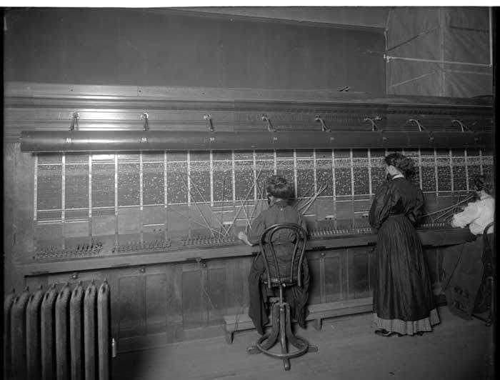 Black-and-white photograph of women operators working at a long wall-mounted telephone switchboard criss-crossed with cords.