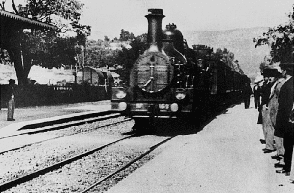 Black-and-white film frame of a steam locomotive arriving at a station platform, seen at an angle, with people waiting on the platform.