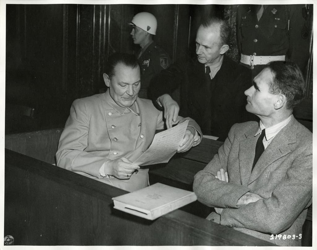 Black-and-white photo of Nazi defendants Göring, Dönitz and Hess conferring with each other in the Nuremberg courtroom dock.