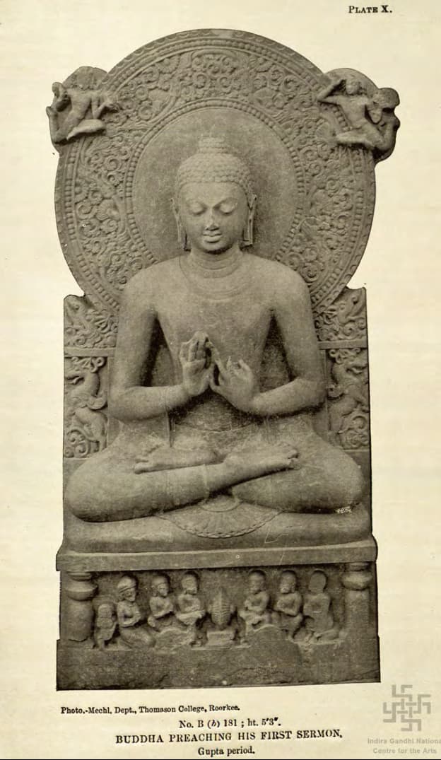 An ancient stone sculpture of the Buddha seated cross-legged with a large carved halo behind his head, his hands raised before his chest in a teaching gesture.
