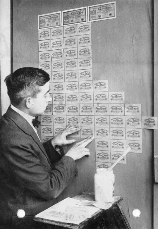 Black-and-white photo of a man in a bow tie pasting rows of German banknotes onto a wall like wallpaper, with a pot of paste beside him.