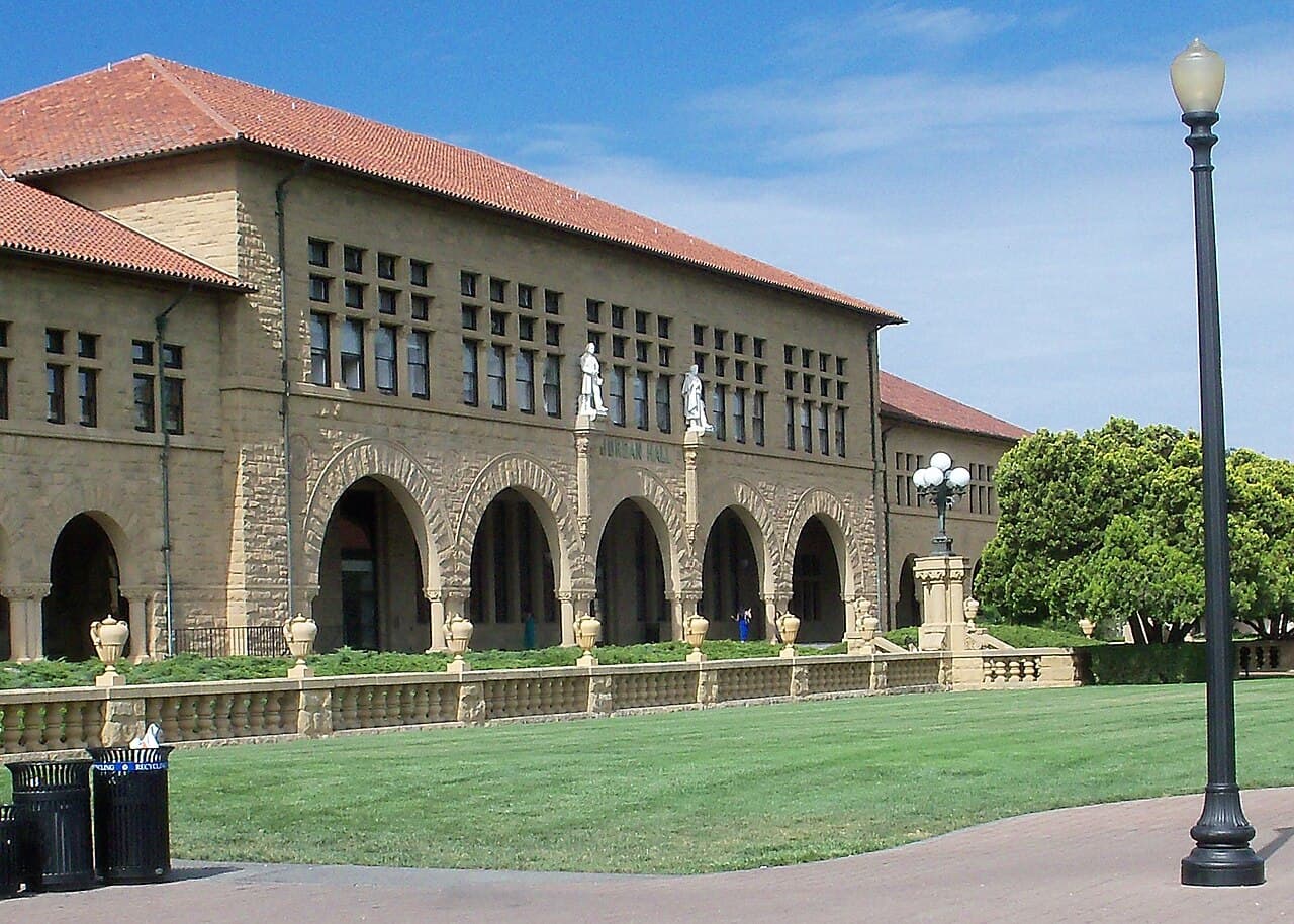 A sandstone university building with arched windows, two statues on its facade and a stone balustrade in front.