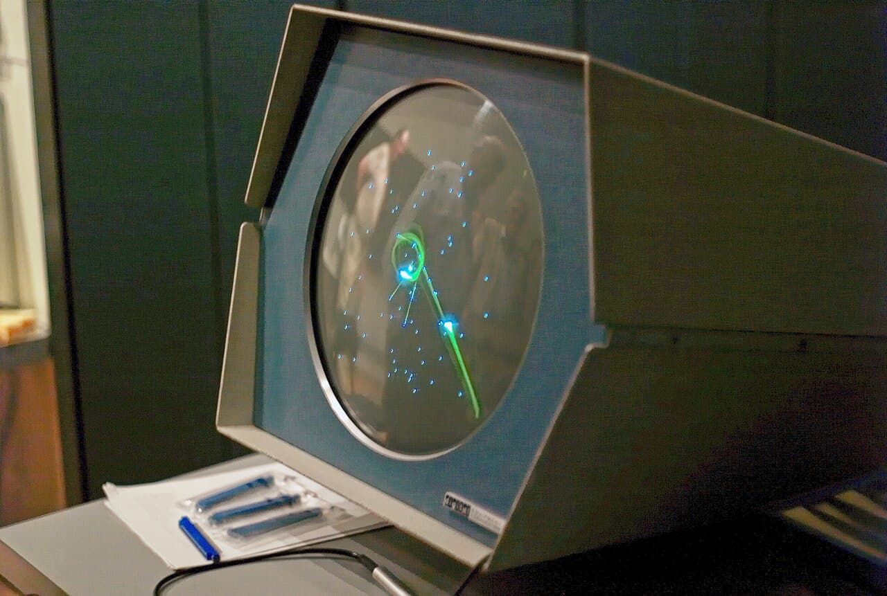 A round computer display showing two small spaceships and a central star, with the PDP-1 console around it