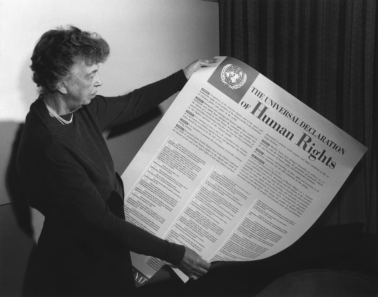 Black-and-white photo of Eleanor Roosevelt holding up a large poster printed with the text of the Universal Declaration of Human Rights.