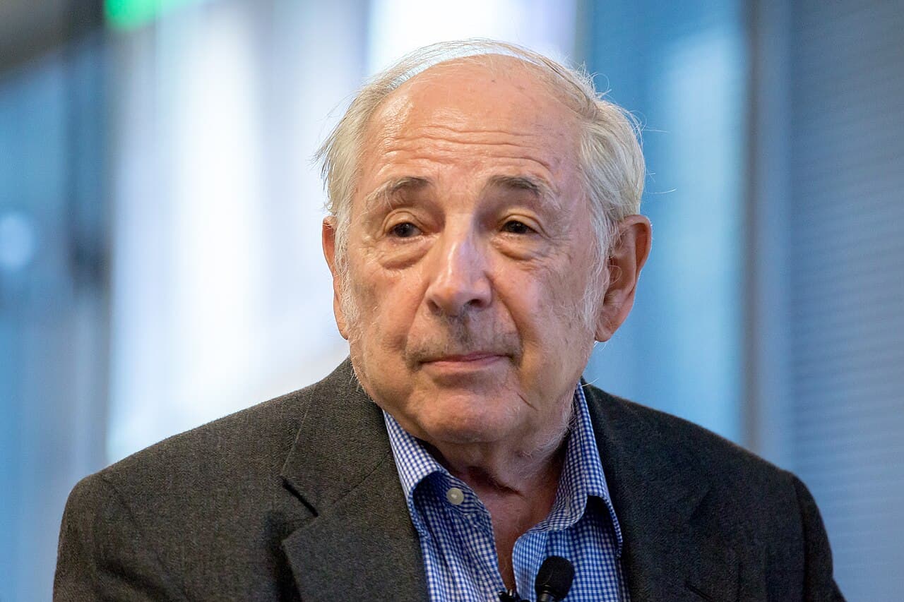 John Searle, an elderly man in a jacket, speaking at a lectern in front of an audience.