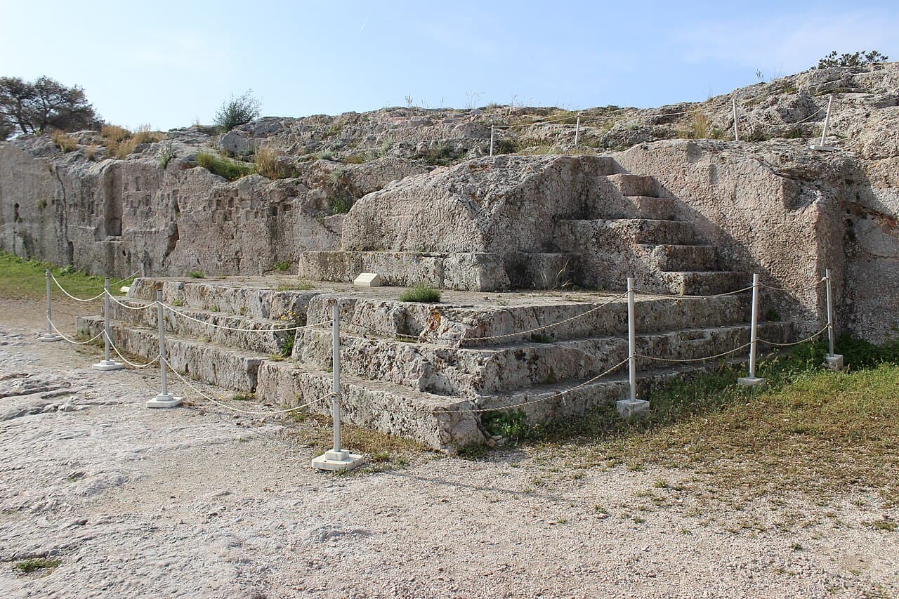 A large stepped block cut into the rock of a hillside in Athens, with trees and the city in the background.