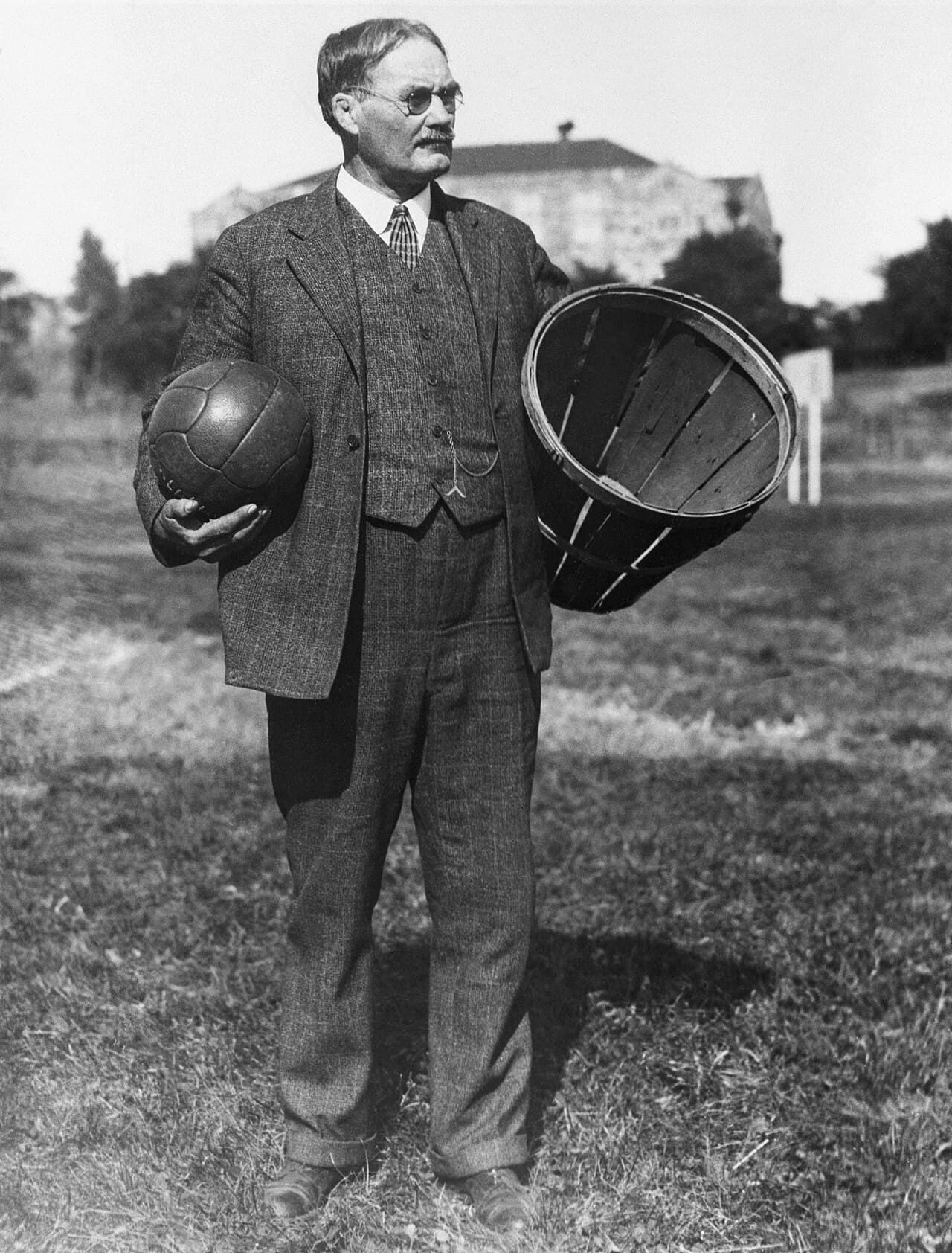Old photograph of James Naismith holding a soccer ball next to a peach basket