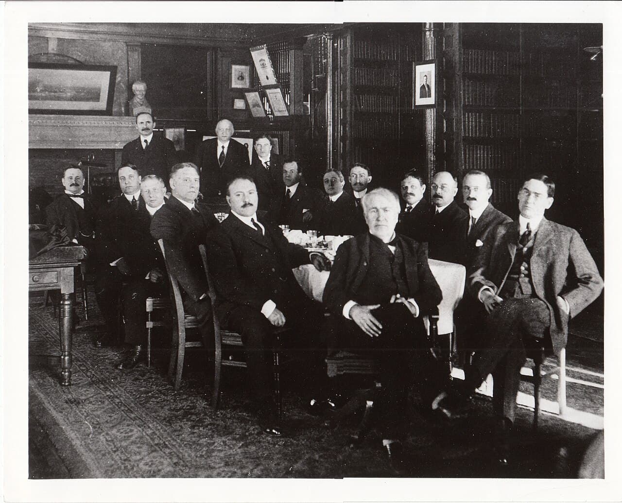 A group of men in dark suits posing indoors around Thomas Edison, in a black-and-white photograph.