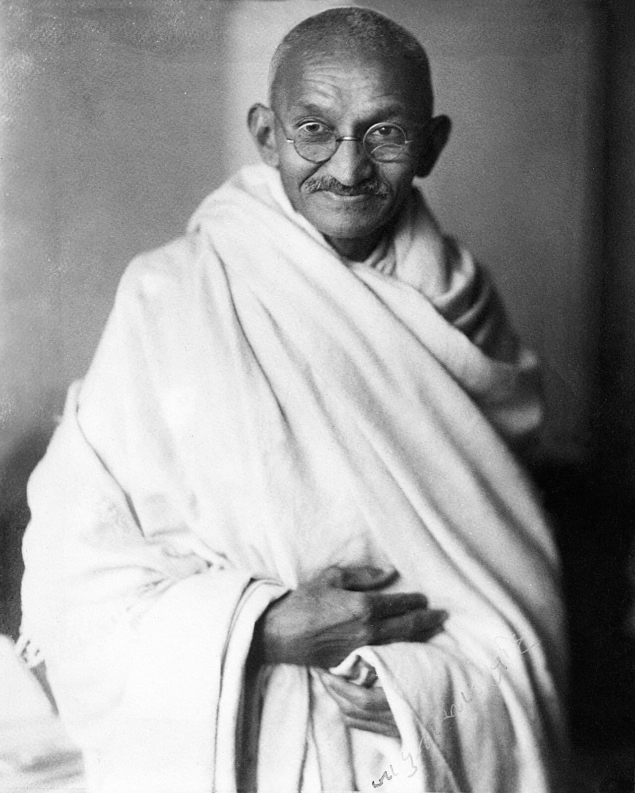 Black-and-white studio portrait of Mahatma Gandhi, smiling, wearing round glasses and a white shawl.
