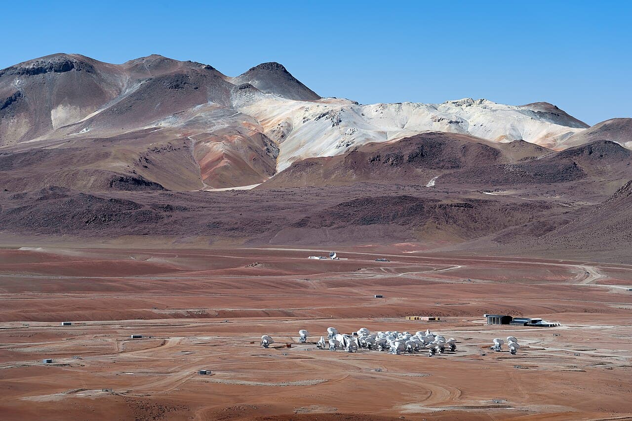 Large white radio telescope dishes of the Atacama Large Millimeter Array standing in the barren, reddish-brown landscape of the Atacama Desert.