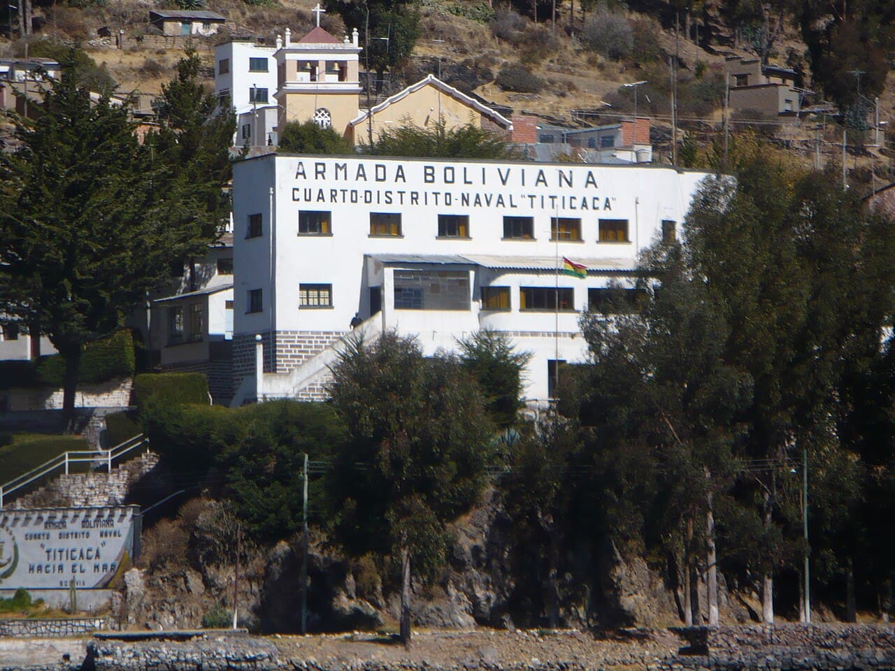 A white building on a rocky lakeshore hillside with large lettering reading 'Armada Boliviana, Cuarto Distrito Naval Titicaca', with a Bolivian flag on the facade.