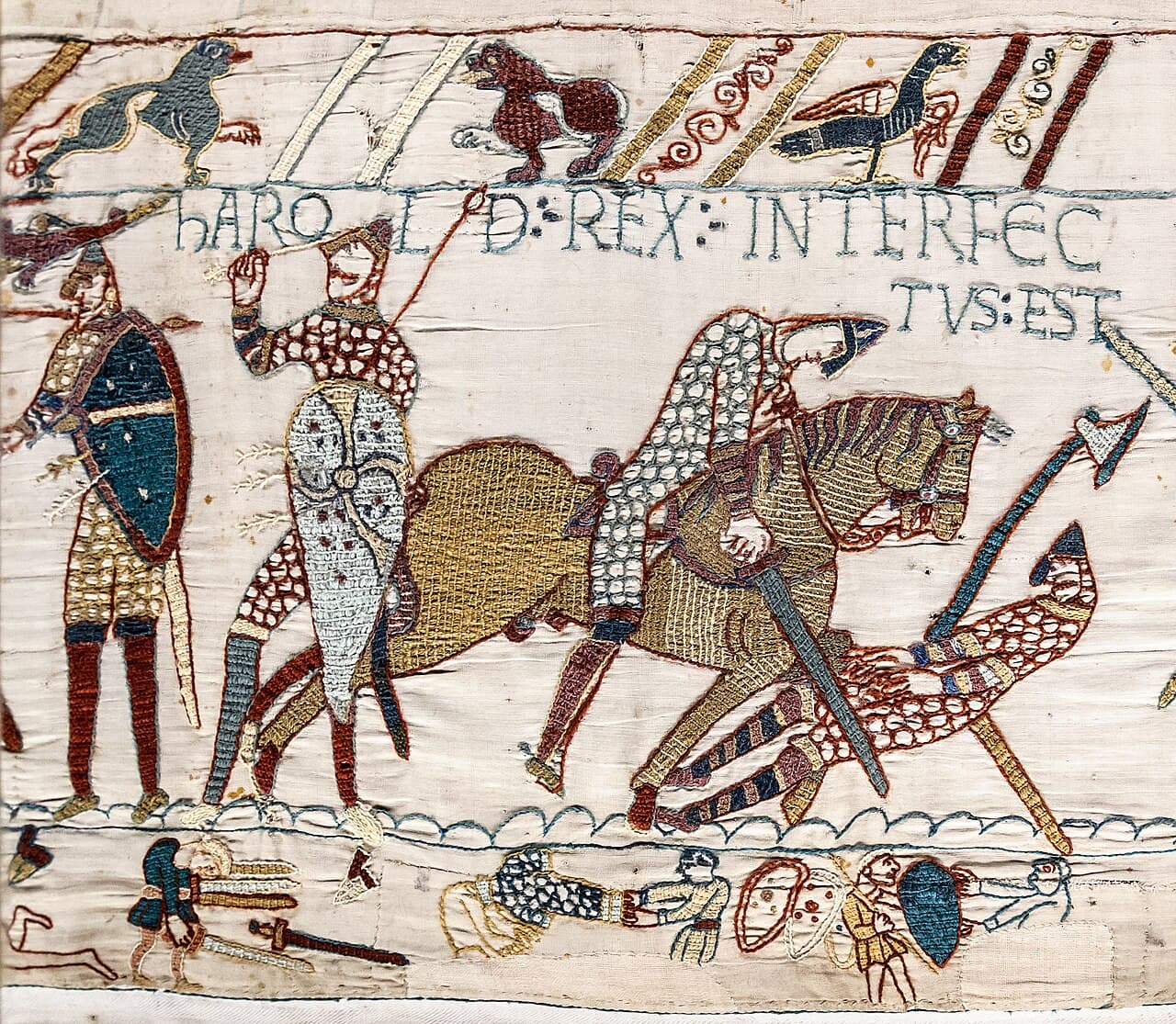 Embroidered scene from the Bayeux Tapestry showing mounted Norman knights and English soldiers fighting, with a Latin inscription above.