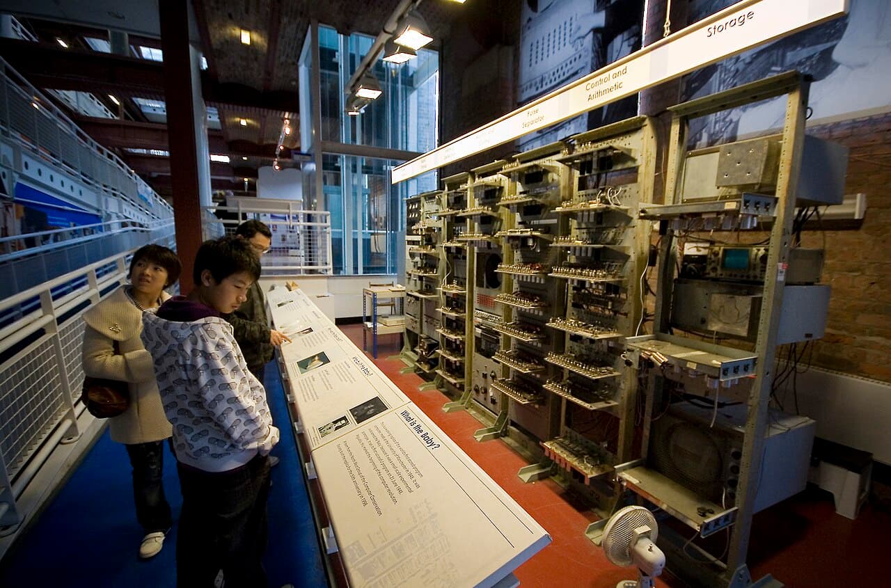 A large metal cabinet covered in racks of vacuum tubes and wiring, a reconstruction of the Manchester Baby computer, with a cathode-ray tube screen visible.
