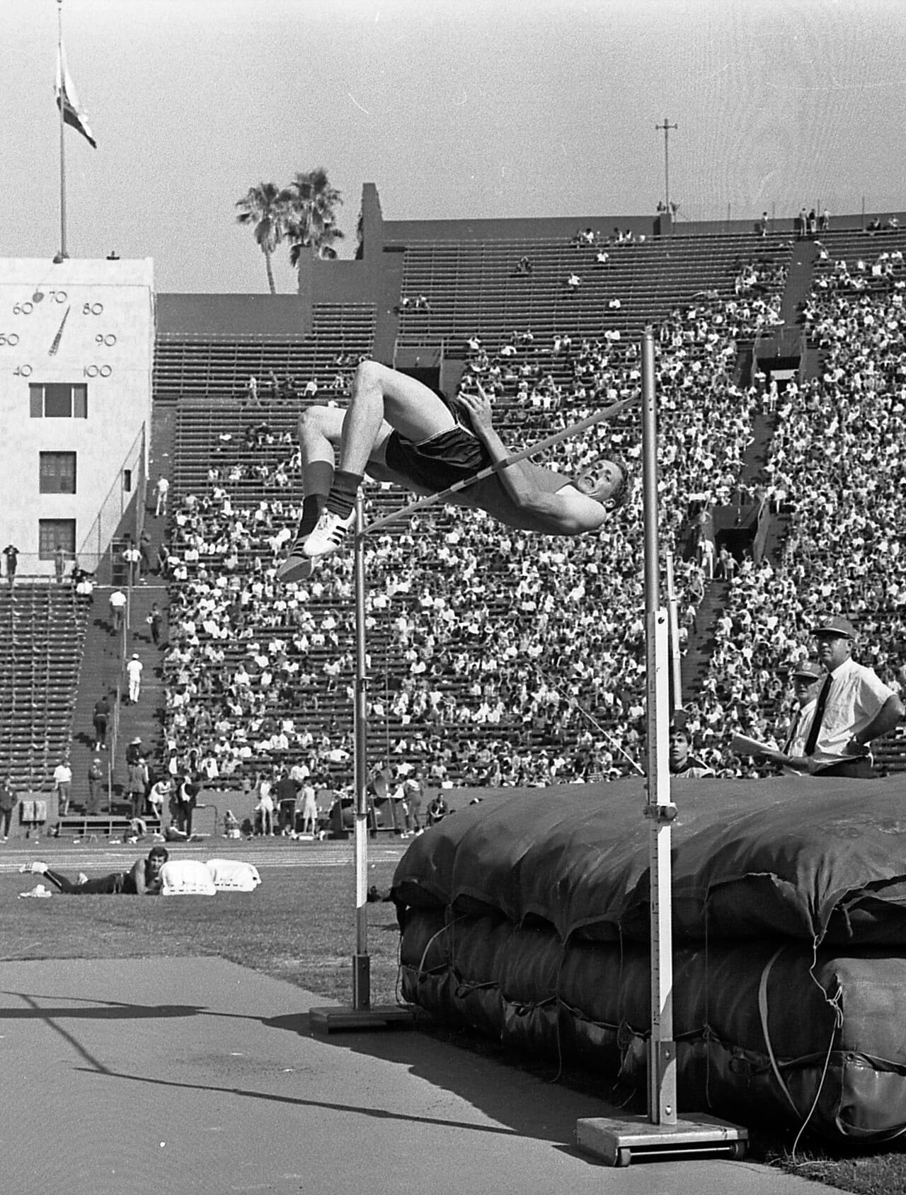 Black-and-white photo of Dick Fosbury arching backwards over a high-jump bar, head and shoulders already past it