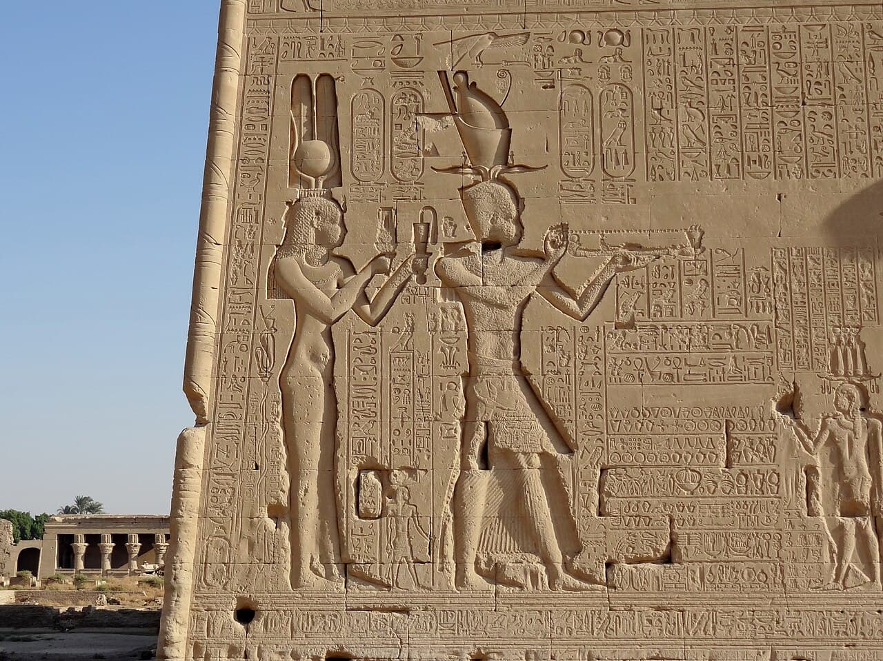A large carved stone relief on a temple wall showing two figures in Egyptian royal crowns and robes, in profile, making offerings to Egyptian gods.