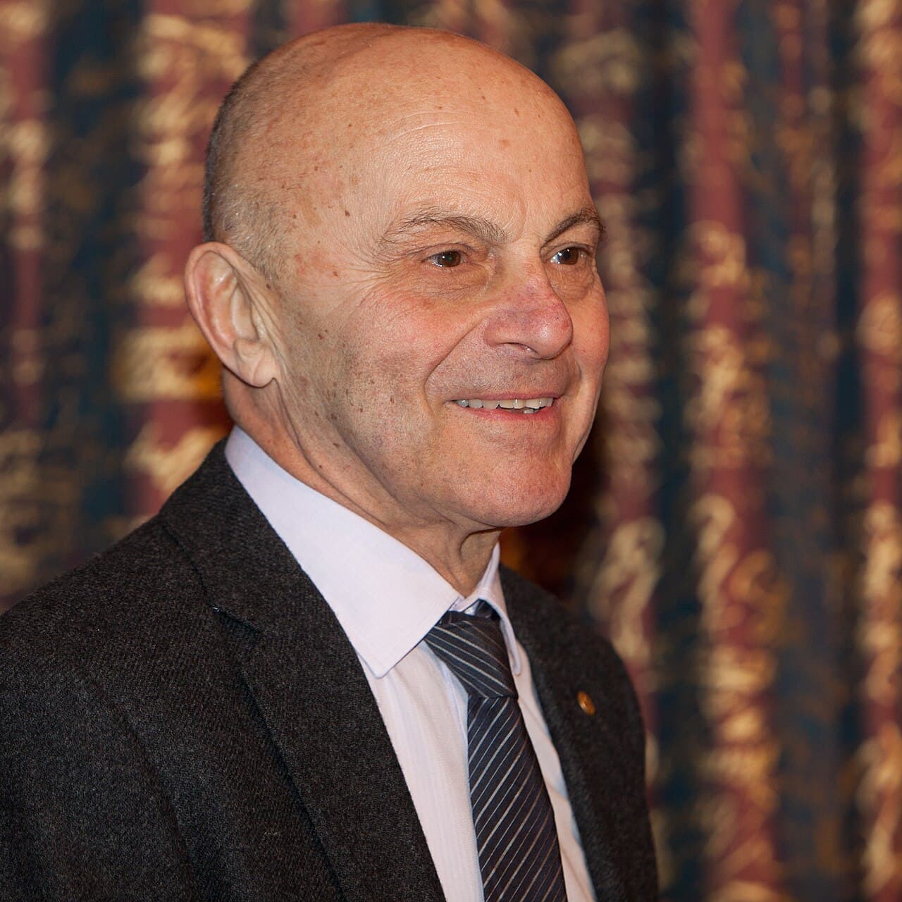 Eugene Fama, an older man in a dark suit, speaking at a press conference table with a microphone.